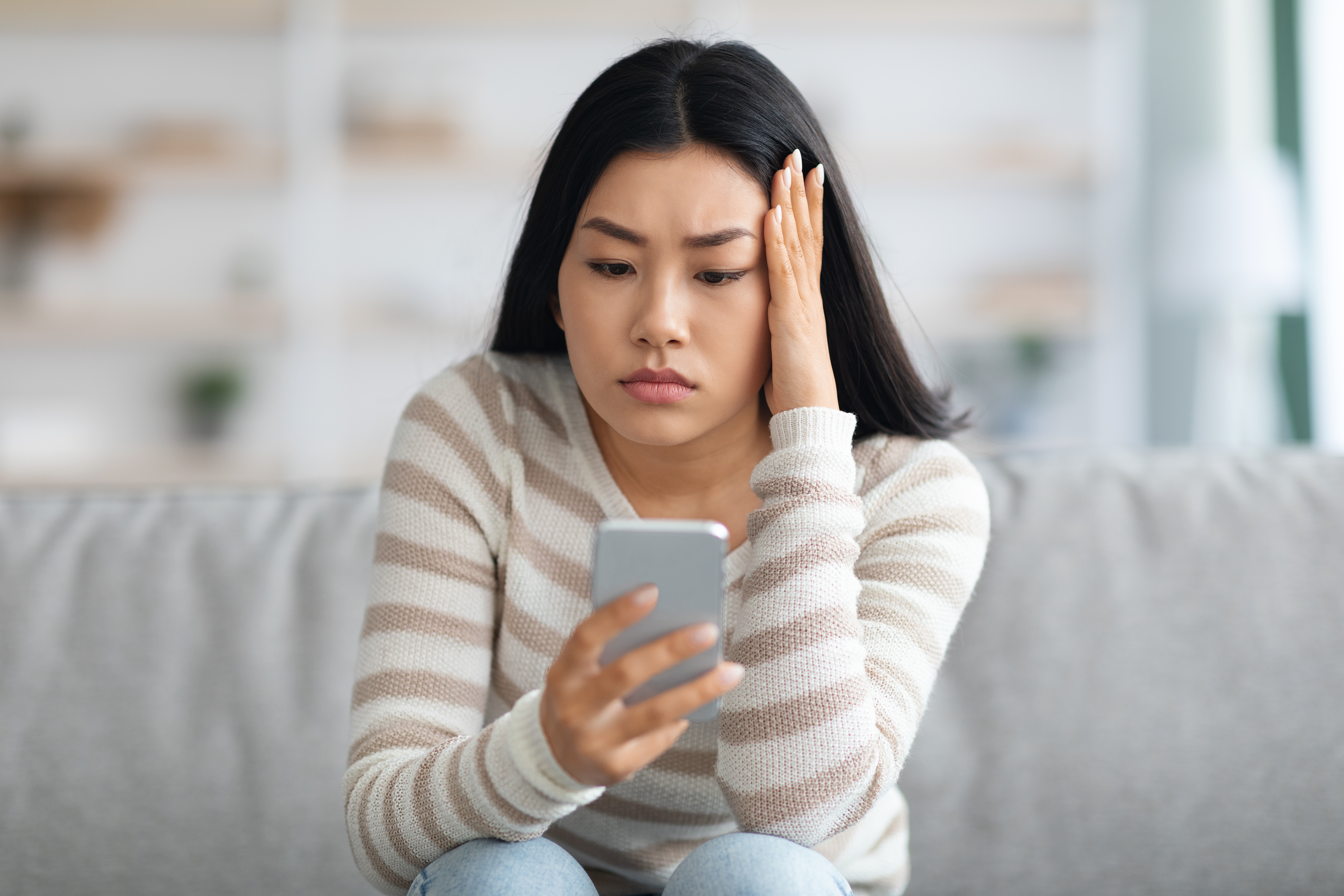 Distressed Asian woman on a couch, holding a smartphone with a worried expression, receiving bad news on the screen.